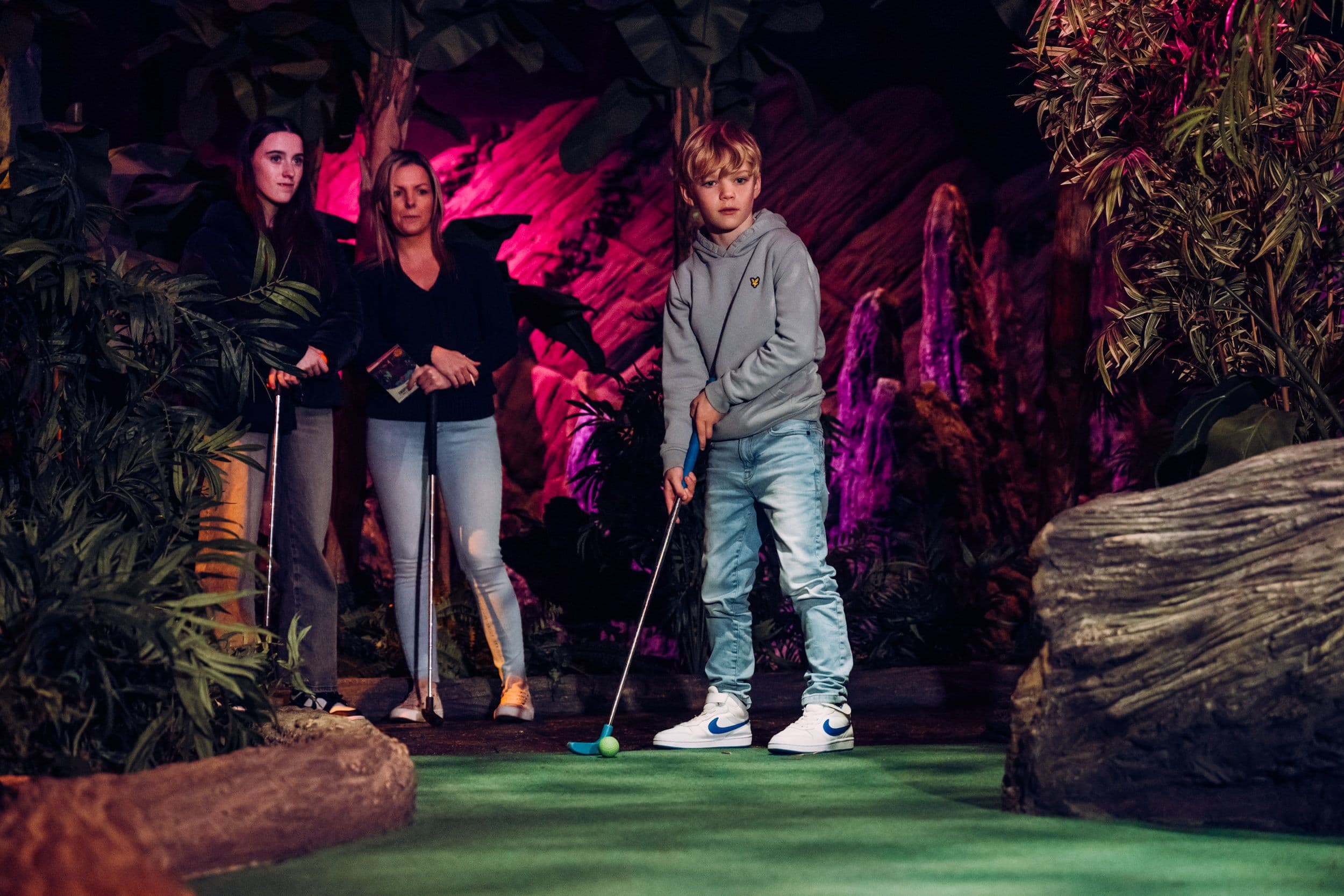 A young teenager putting at Treetop Golf while two women watch.